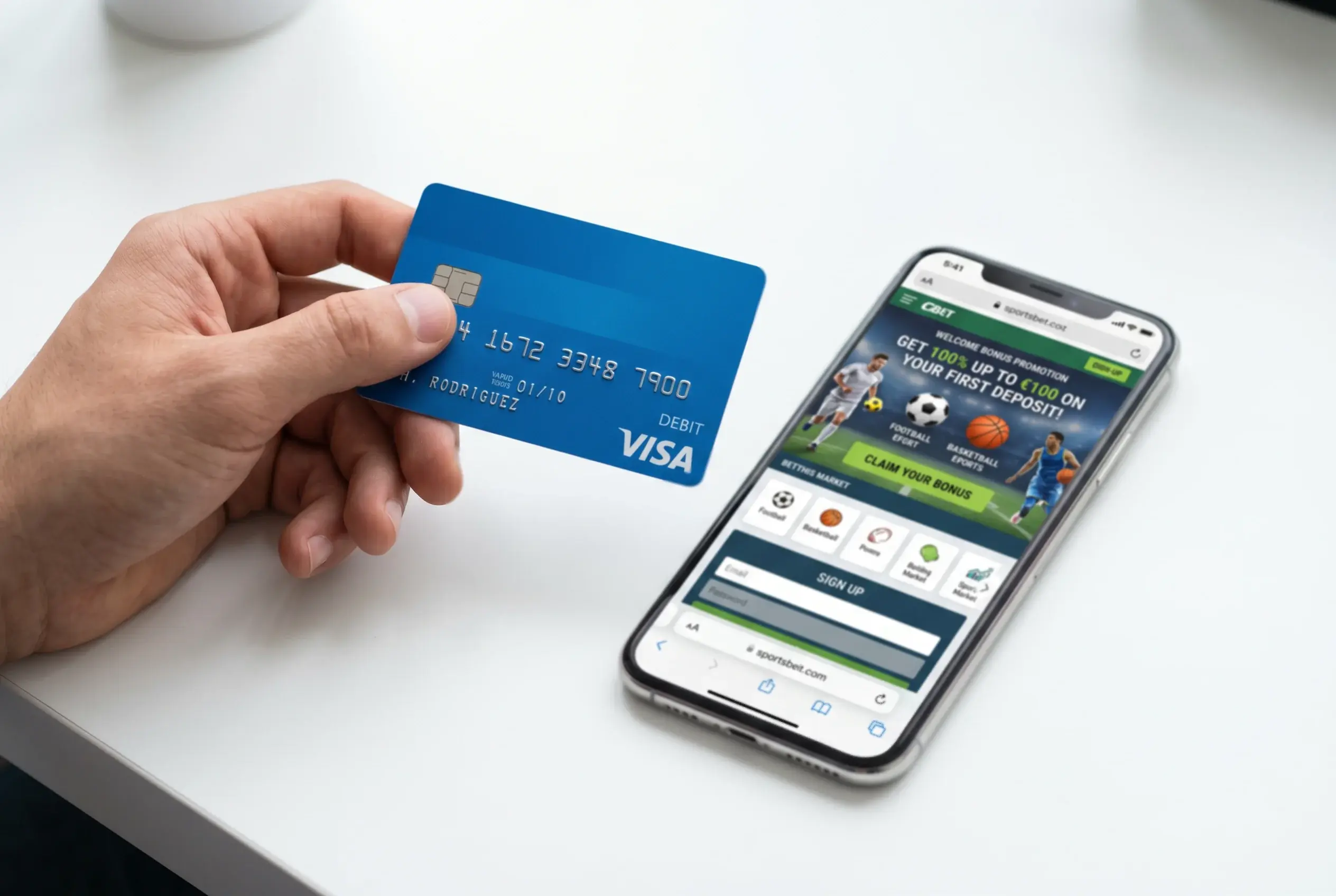 Mano sosteniendo una tarjeta Visa de débito junto a una pantalla de móvil que muestra una promoción de apuestas deportivas